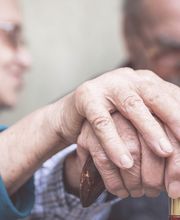 Due anziani si tengono per mano sopra un bastone da passeggio, in un gesto di affetto.