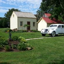 Vintage white car parked on grass near small, quaint houses with garden and trees.