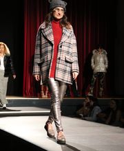 Model auf dem Laufsteg in kariertem Mantel, rotem Pullover und silbernen Hosen vor rotem Vorhang.