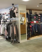 Kleidungsgeschäft mit Regalen voller Jeans, Hemden und Jacken; umkleidekabine im Hintergrund.