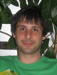 Ein Mann in einem grünen Shirt lächelt leicht vor einer Pflanze im Hintergrund.