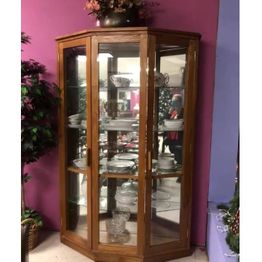 Wooden corner cabinet with glass doors displaying plates and glassware against a pink wall.