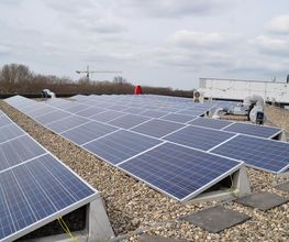 Paneles solares en un techo plano, cielo parcialmente nublado, grúa y árboles al fondo.