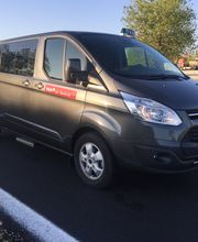 Furgoneta gris en una carretera asfaltada con árboles al fondo y un número de taxi en el parabrisas.