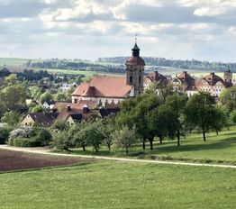 Kleines Dorf mit Kirche und historischen Gebäuden, umgeben von grünen Feldern und Bäumen.