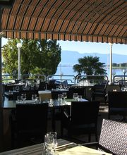 Überdachte Terrasse mit Tischen und Stühlen, Blick auf See und Berge im Hintergrund.