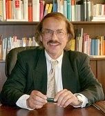 Mann im Anzug mit Brille lächelt, sitzt vor einem Bücherregal.