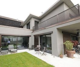 Modernes Haus mit Terrasse, Balkon, Rasen und Pflanzen, graue Fassade und Holzverkleidung.