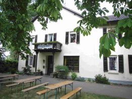 Weiße, zweistöckige Villa mit grünen Fensterläden, Biertische im Vordergrund, Bäume umgeben das Haus.