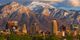 City skyline with tall buildings in front of snow-covered mountains under a cloudy sky.