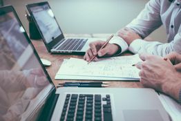 Due persone lavorano al computer con documenti e appunti su una scrivania.
