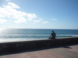 Person sitzt auf einer Mauer und blickt aufs Meer unter klarem Himmel.