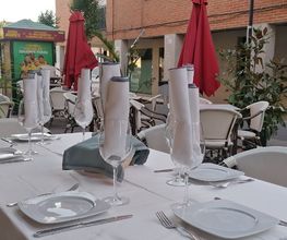 Mesa exterior de restaurante con manteles blancos y copas, rodeada de sillas y sombrillas rojas.