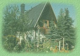 Grünes Landhaus mit spitzem Dach, umgeben von Bäumen und buntem Garten im Vordergrund.