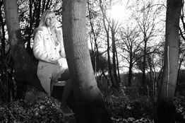 Frau in weißem Mantel sitzt auf einem Baum, im Hintergrund Wald und Sonnenschein.