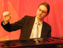 Ein Mann im Anzug spielt Klavier vor einem roten Hintergrund.