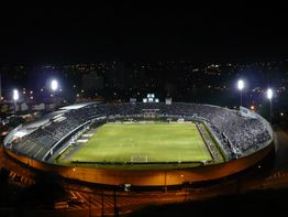 Estádio de futebol iluminado à noite com torcida e cidade ao fundo.