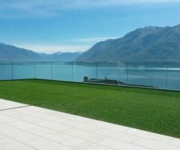Terrasse mit Glasgeländer, Blick auf See und Berge unter blauem Himmel.