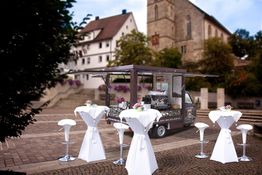 Im Bild steht ein mobiler Kaffeestand mit Stehtischen auf einem gepflasterten Platz vor alten Gebäuden.