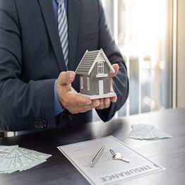 Man holding house model over insurance papers on desk with cash and keys.