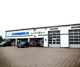 Mehrere Autos vor einer Autowerkstatt mit Glasgaragentoren und einem großen Firmenlogo.