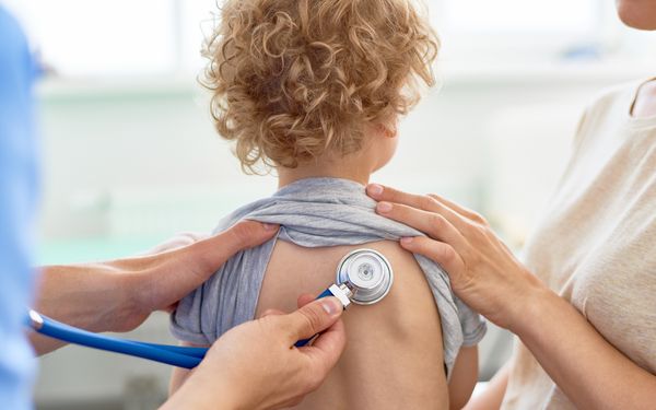 Ein Kind sitzt mit dem Rücken zum Bild. Ein Arzt hört sich die Luge des Patienten mithilfe eines Stetoskops an.