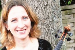 Frau lächelt vor einem Baum, eine Violine ist im Bild sichtbar, sommerlicher Hintergrund.
