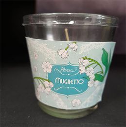 Candela profumata in bicchiere di vetro, etichetta con fiori di mughetto su sfondo azzurro.