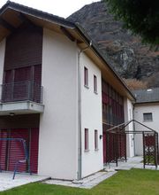 Modernes zweistöckiges Haus mit braunen Fensterläden vor einem felsigen Hügel und Rasenfläche.