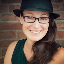 Eine Frau mit Brille und Hut lächelt vor einer Backsteinwand.