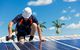 Worker in helmet installing solar panels on a sunny rooftop with palm trees in the background.