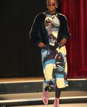 Person in bunter Mode auf Laufsteg, trägt Sonnenbrille und rosa Schuhe, schwarzer Hintergrund.