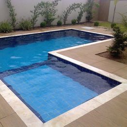 Piscina retangular com ladrilhos azuis, cercada por piso de madeira clara e plantas ao fundo.