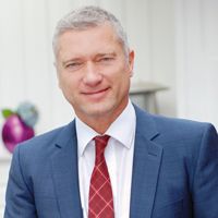 Ein Mann im Anzug mit roter Krawatte lächelt vor einem verschwommenen Hintergrund.