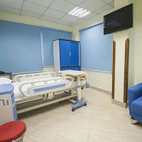 Hospital room with a bed, blue chair, TV, and blue blinds.
