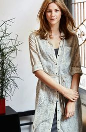 Vrouw met los haar draagt een lichtgekleurde jurk en staat naast een plant in een interieur.