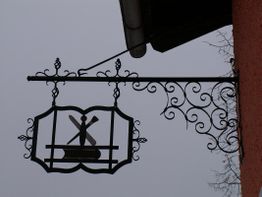 Schmiedeeisernes Wirtshausschild mit einem stilisierten Mühlensymbol, vor bewölktem Himmel.