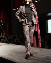 Person im karierten Anzug auf Laufsteg, Mantel über der Schulter, lächelt und posiert selbstbewusst.