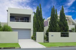 Modern white house with a balcony, tall trees, and a manicured lawn under a blue sky.