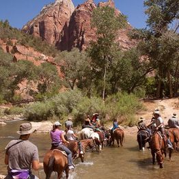 People riding horses through a shallow river with rocky cliffs and trees in the background.