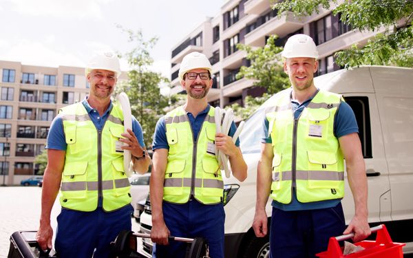 Drei Mechaniker mit Warnwesten, tragen ihr Werkzeug in der Hand und lächeln in die Kamera.