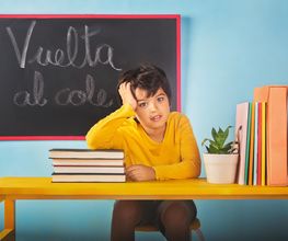 Niño en clase con expresión pensativa, pizarra dice "Vuelta al cole", libros y planta en mesa.
