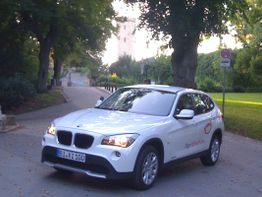 Weißer BMW SUV auf einem Parkweg mit Bäumen und einem Gebäude im Hintergrund.