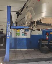 Carro prata elevado em plataforma de oficina mecânica. Sem roda traseira. Ferramentas ao fundo.