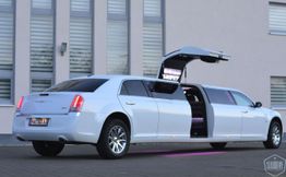 Weiße Stretchlimousine mit geöffneter Flügeltür vor einem Gebäude mit großen Fenstern.