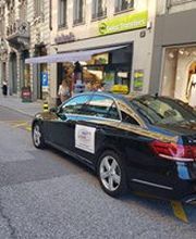 Schwarzes Auto parkt auf einer Stadtstraße vor Geschäften mit Passanten im Hintergrund.