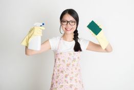 Woman with glasses, apron, and gloves holding a spray bottle and sponge, smiling.