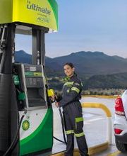 Mujer en estación de servicio BP, sosteniendo manguera de gasolina junto a un coche blanco.