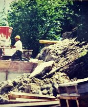 Bauarbeiter auf Baustelle mit Betonmischer und Erdhaufen im Hintergrund, umgeben von Bäumen.