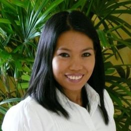 Eine lächelnde Frau mit dunklem Haar vor grünen Pflanzen.